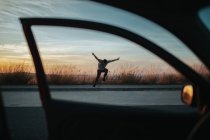 From car view of full body young man in casual wear jumping on skateboard while performing kickflip on asphalt road against dusky sky - foto de stock