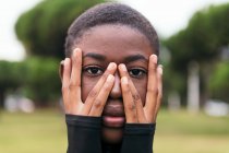 Joven hembra afroamericana tierna con la cara cubierta de pelo corto mientras mira a la cámara en la ciudad en el día de verano - foto de stock