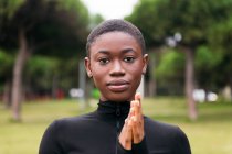 Jovem macio étnico feminino no preto vestuário com cabelo curto olhando para câmera no gramado urbano no verão — Fotografia de Stock