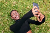 Молода етнічна жінка в повсякденному одязі з бездротовим гарнітуром взяла автопортрет на мобільний телефон, лежачи в парку — стокове фото