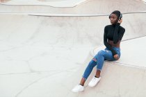 Seitenansicht einer jungen, nachdenklichen Afroamerikanerin in zerrissenen Jeans, die Musik aus drahtlosen Kopfhörern hört, während sie nach vorne blickt — Stockfoto