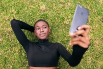 Young ethnic female in casual apparel with wireless headset taking self portrait on mobile phone lying in a park — Stock Photo