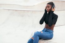 Seitenansicht einer jungen, nachdenklichen Afroamerikanerin in zerrissenen Jeans, die Musik aus drahtlosen Kopfhörern hört, während sie nach vorne blickt — Stockfoto