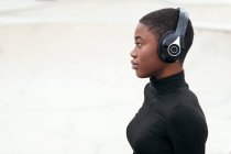 Vista lateral de una joven afroamericana contemplativa en jeans rasgados escuchando música de auriculares inalámbricos mientras mira hacia adelante - foto de stock