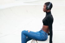 Вид сбоку на молодую созерцательную афроамериканку в разорванных джинсах, слушающую музыку из беспроводных наушников, глядя вперед — стоковое фото