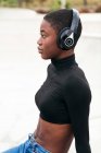 Side view of young contemplative African American female in ripped jeans listening to music from wireless headphones while looking forward — Stock Photo