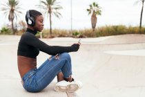 Young glad ethnic female in wireless headphones surfing internet on cellphone while listening to song in city — Stock Photo