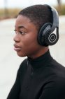 Сторона зору молодої споглядальної афроамериканської жінки в пошматованих джинсах, яка слухає музику з бездротових навушників, дивлячись вперед. — стокове фото