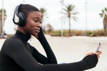 Young glad ethnic female in wireless headphones surfing internet on cellphone while listening to song in city — Stock Photo