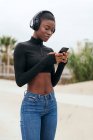 Niedriger Winkel junger fröhlicher ethnischer Frauen in drahtlosen Kopfhörern, die auf dem Handy im Internet surfen, während sie in der Stadt Lieder hören — Stockfoto