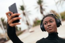 Молодая этническая женщина в повседневной одежде с беспроводной гарнитурой делает автопортрет на мобильном телефоне в парке — стоковое фото