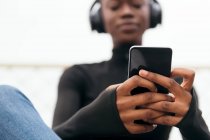 Низький кут молодої радий етнічної жінки в бездротових навушниках, що серфінгують Інтернет на мобільному телефоні, слухаючи пісню в місті — стокове фото