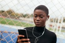 Mensajería de texto femenina étnica enfocada a los jóvenes en el teléfono celular mientras escuchan música contra la valla de la red en la ciudad - foto de stock