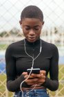 Mensajería de texto femenina étnica enfocada a los jóvenes en el teléfono celular mientras escuchan música contra la valla de la red en la ciudad - foto de stock
