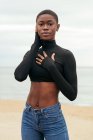 Joven hembra afroamericana suave con ropa casual tocando la mejilla mientras mira la cámara en la costa del océano - foto de stock