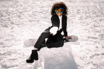 Cheerful African American female athlete in trendy wear and protective glasses sitting on snowboard while looking at camera in winter — Stock Photo