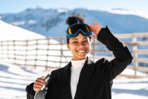 Щаслива етнічна спортсменка в стильному одязі та захисних окулярах, що несуть сноуборд на засніженій гонці на сонячному світлі, дивлячись на камеру — стокове фото