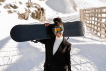 Happy ethnic sportswoman in stylish wear and protective glasses carrying snowboard on snowy mount in sunlight looking at camera — Stock Photo