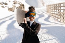 Glückliche ethnische Sportlerin in stylischer Kleidung und Schutzbrille mit Snowboard auf schneebedecktem Berg im Sonnenlicht und Blick in die Kamera — Stockfoto