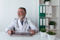 Мечтательный пожилой врач в очках со стетоскопом, смотрящий на стол с планшетом в больнице — стоковое фото