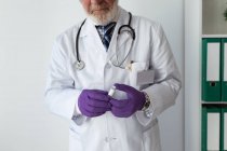 Crop anziano barbuto medico maschio in uniforme e guanti monouso dimostrando piccola bottiglia con sostanza liquida blu su sfondo bianco — Foto stock