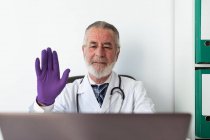 Médico senior con guantes uniformados y estériles mostrando gesto de saludo contra netbook durante una videollamada en el hospital - foto de stock