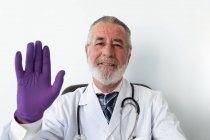 Médico senior con guantes uniformados y estériles mostrando gesto de saludo contra netbook durante una videollamada en el hospital - foto de stock