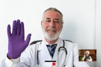 Senior male doctor with in uniform and sterile gloves showing greeting gesture against netbook during video call in hospital — Stock Photo