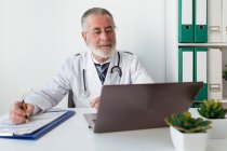 Médico senior en uniforme escribiendo notas en papel durante la consulta de salud en línea en el ordenador portátil en el hospital - foto de stock