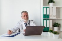 Старший врач мужского пола в униформе пишет заметки на бумаге во время онлайн консультации по вопросам здоровья на портативном компьютере в больнице — стоковое фото