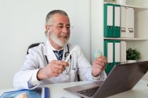Hombre mayor ortodoncista mostrando cepillo de dientes y hilo dental mientras habla contra netbook durante el chat de vídeo en la mesa en el hospital - foto de stock