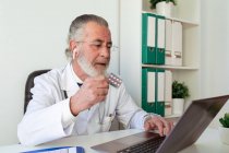 Elderly male doctor in earbud showing medications while talking on video chat against netbook during online health consultation in hospital — Stock Photo