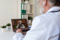 Visão traseira da cultura anônimo idoso médico masculino em fone de ouvido sem fio falando com mulher em chamada de vídeo no netbook na clínica — Fotografia de Stock