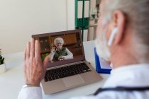Back view of crop anonymous elderly male doctor in wireless earbud speaking with woman on video call on netbook in clinic — Stock Photo