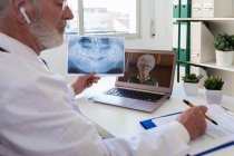 Vista laterale di un anziano medico maschio con auricolare senza fili che parla con una donna in videochiamata su netbook in clinica — Foto stock