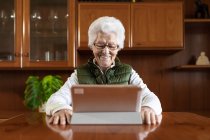 Friendly elderly female showing toothy smile against tablet while video chatting in house — Stock Photo