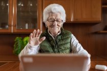 Friendly elderly female in wireless earbuds showing greeting gesture against tablet while video chatting in house — Stock Photo