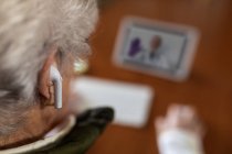 Vue de dessus du dos d'une patiente âgée anonyme de culture dans des écouteurs TWS parlant avec un médecin sur une tablette pendant un appel vidéo à la maison — Photo de stock