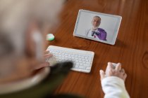 Vue de dessus du dos d'une patiente âgée anonyme de culture dans des écouteurs TWS parlant avec un médecin sur une tablette pendant un appel vidéo à la maison — Photo de stock
