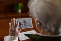 Сверху спины просматривается анонимная пожилая женщина в наушниках TWS, разговаривающая с врачом на планшете во время видеозвонка домой — стоковое фото