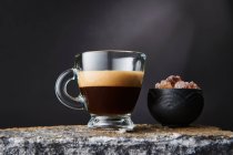 Прозорий келих міцної чорної кави з піною біля миски коричневих кубиків цукру на чорному тлі — стокове фото