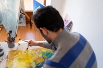 Неузнаваемый мужчина-художник, использующий профессиональную кисть в процессе покраски на картонном листе рядом с художественными инструментами в мастерской — стоковое фото