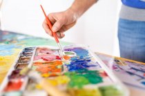 Crop anonymous male painter mixing paints with brush using watercolor palette while working in art studio — Stock Photo