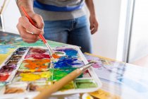 Crop anonymous male painter mixing paints with brush using watercolor palette while working in art studio — Stock Photo