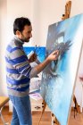Side view of young bearded ethnic painter in casual wear painting with brush on canvas in art studio - foto de stock