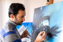 Side view of young bearded ethnic painter in casual wear painting with brush on canvas in art studio — Stock Photo