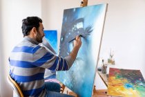 Side view of young bearded ethnic painter in casual wear siting in a chair painting with brush on canvas in art studio - foto de stock