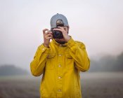 Giovane fotografo maschio in giacca di denim giallo scattare foto sulla macchina fotografica mentre in piedi su nebbioso sfondo della natura offuscata — Foto stock