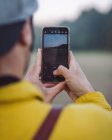 Back view young male backpacker in yellow denim jacket taking photo on smartphone while standing on foggy meadow in nature — Fotografia de Stock