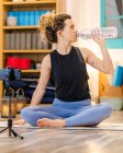 Спокійна жінка з кучерявим волоссям у спортивному одязі, що сидить на підлозі та п'є пляшку води під час перерви в онлайн-сесії йоги в студії — стокове фото